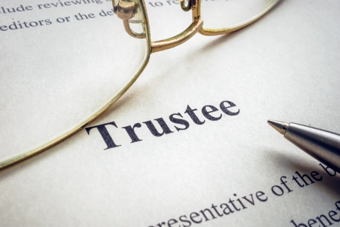 A piece of paper displays the word Trustee above a pen and underneath a pair of gold-rimmed glasses.