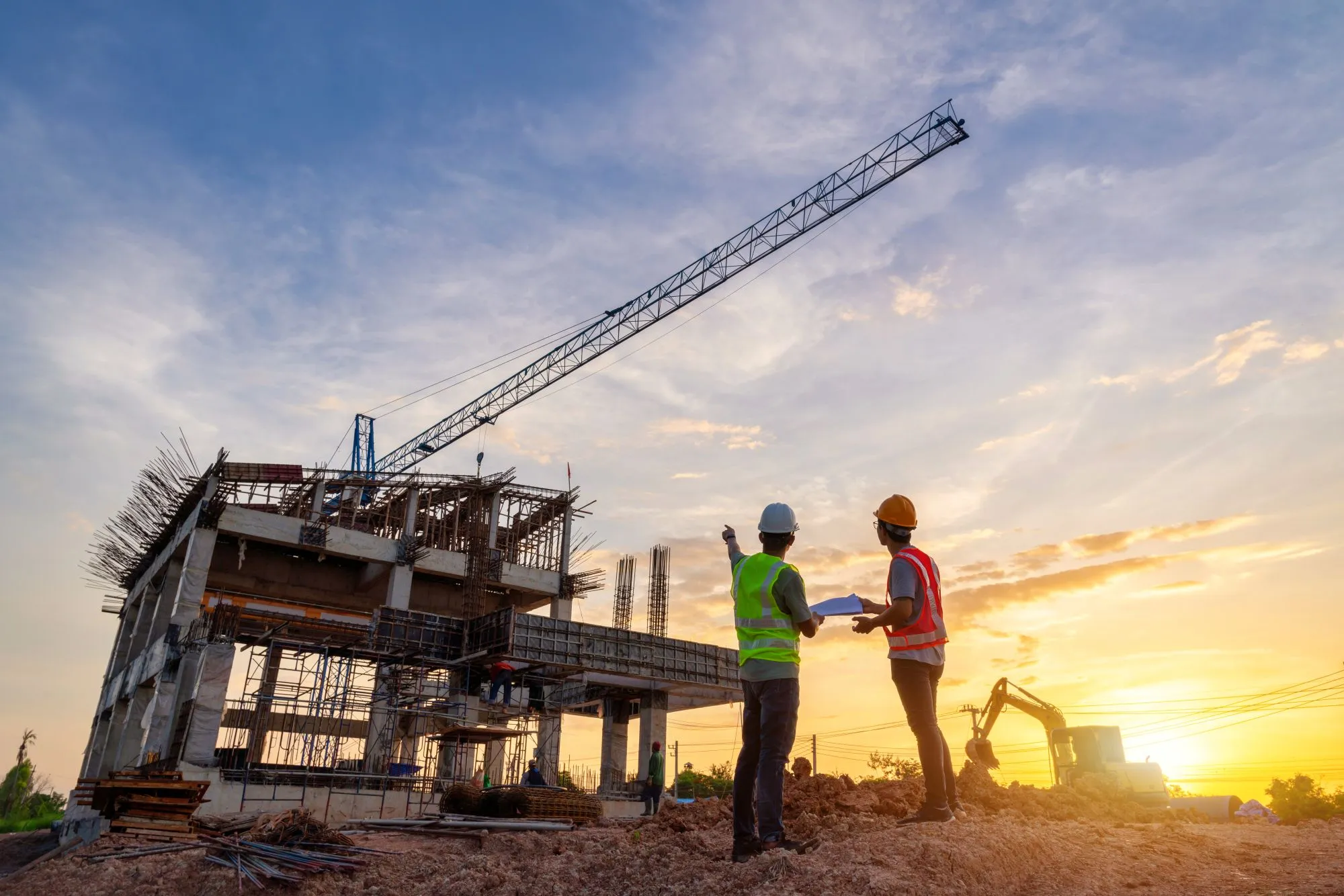 Two people in hi-vis vests and hardhats looking at a commercial building being constructed at sunset. 