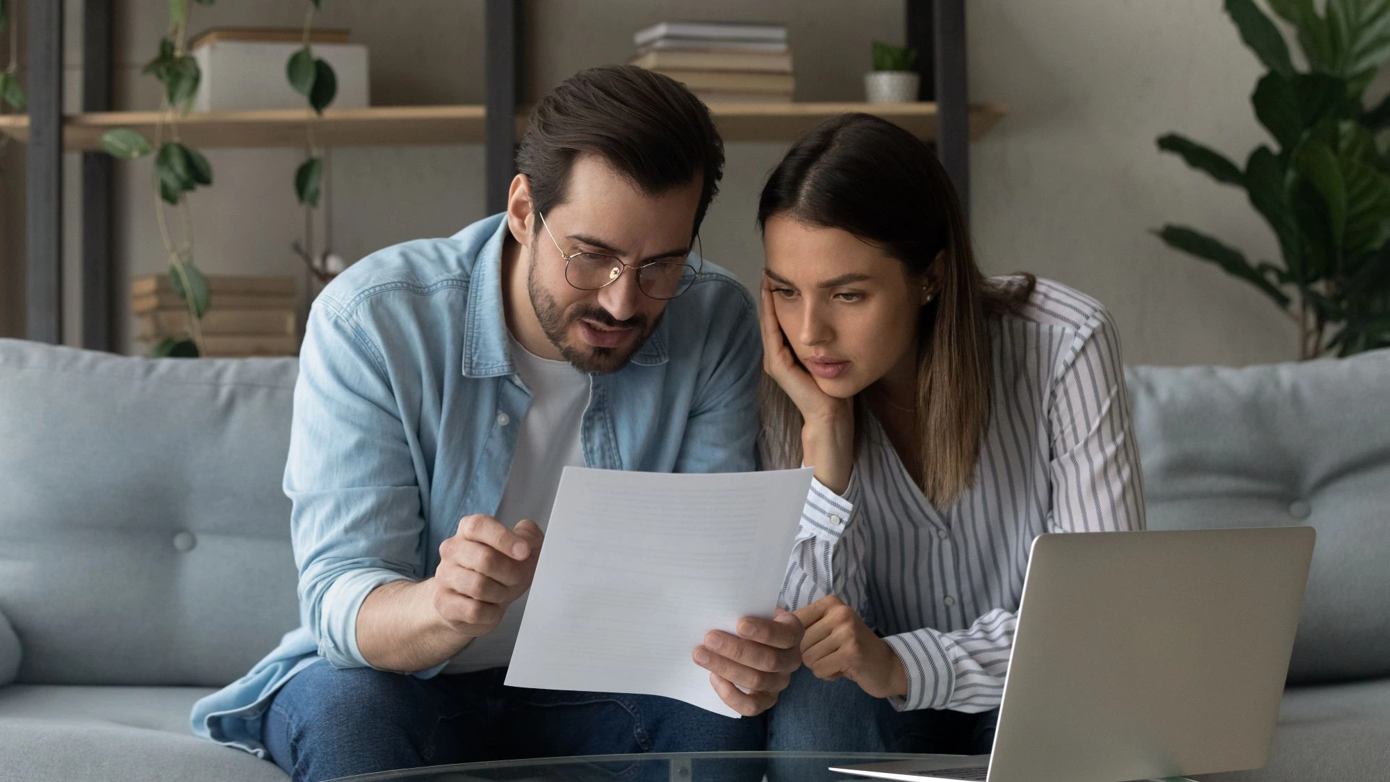 Couple reading over tenancy document sat on grey sofa with a laptop in front of them. 
