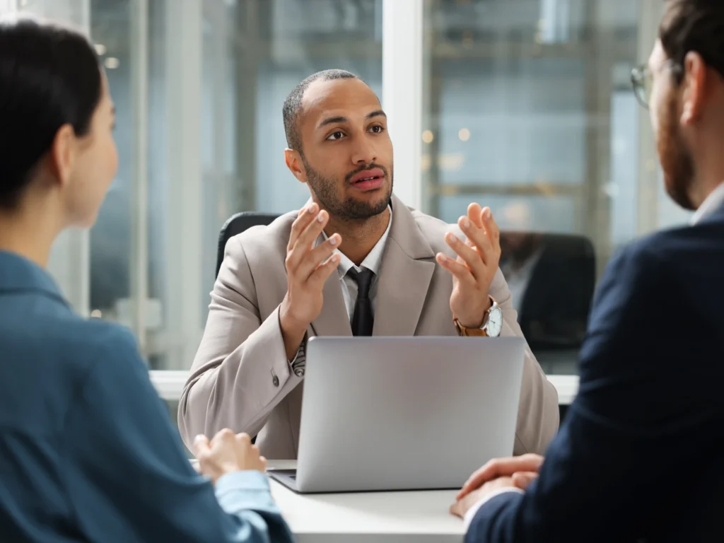 A solicitor in a beige suit sat behind a laptop, speaking with two business owners sat side by side with their hands on the table.